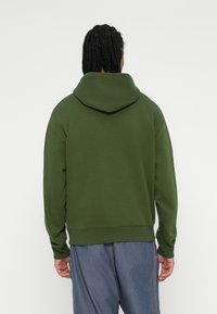 Zelena kapuljača z velikim, prilagodljivim pokrovom, rebrastimi manžetami in robom. Nosi se z ohlapnimi modrimi kavbojkami, ki imajo sproščen dizajn in mehak material.