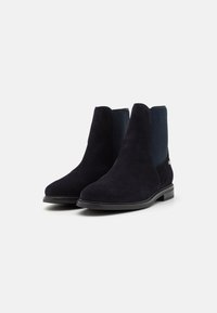 Botas de tobillo de ante en azul oscuro con una textura suave, paneles laterales elásticos y una suela de goma negra. Diseño limpio y minimalista, punta redondeada.