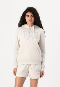Bežinė „Nike“ džemperis su priekinėmis kišenėmis ir raišteliu, papuoštas dideliu „Nike Sportswear“ logotipu. Derinamas su atitinkančiomis bežinėmis šortais.