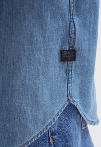 Ourlet d'une chemise en denim bleu clair avec une étiquette noire G-Star RAW sur un jean bleu avec coutures visibles et détail de rivet orange.