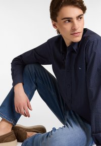 Camicia blu navy con piccoli pois rossi e bianchi, indossata con jeans azzurri e scarpe slip-on marroni, in posa seduta.