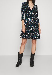 Svart omslagsklänning med v-ringning, 3/4 ärmar och utsvängd kjol. Har färgglada prickmönster i blått, grönt och rosa. Matchas med svarta cowboystövlar.