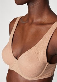 Lace-trimmed, light beige bra with a deep V-neckline, wide straps, and smooth texture, showcasing floral patterns on the fabric.