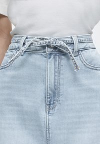 Φούστα από ανοιχτό μπλε τζιν με ζώνη από το ίδιο ύφασμα δεμένη στη μέση, μπροστινές τσέπες και εμφανείς ραφές, συνδυασμένη με λευκή μπλούζα που έχει μπει μέσα στη φούστα.