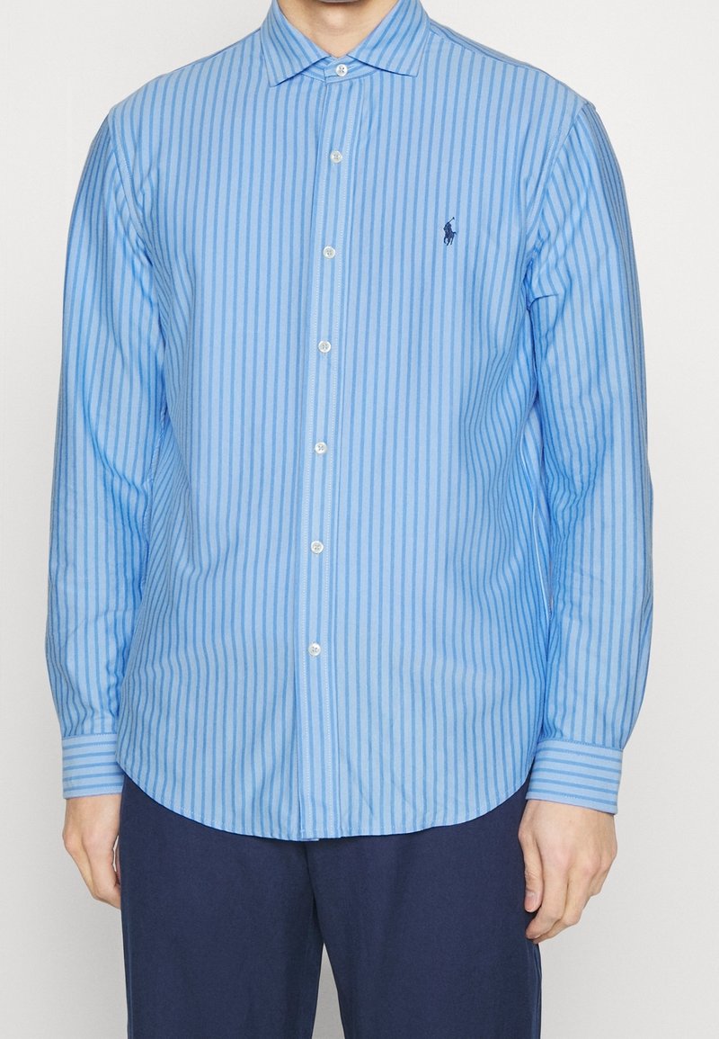 Camicia azzurro chiaro con bottoni, strisce verticali strette, maniche lunghe e un piccolo logo sul petto. Tessuto di cotone con trama liscia.