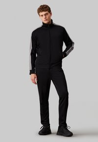 Tuta nera con giacca con zip e spalle grigie, pantaloni abbinati. Il materiale sembra liscio con una vestibilità aderente.