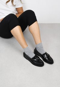Mocasines de cuero negro con un fleco decorativo y detalles de costura, combinados con calcetas de canalé gris claro. Sentados sobre una superficie gris claro.