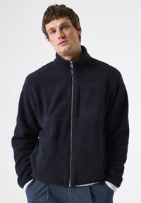 Chaqueta de forro polar azul marino con un cuello alto, cierre completo y un gran bolsillo en el pecho. Presenta una tela texturada y suave, así como puños ajustables.