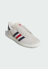 Beige suede sneaker with red and navy stripes, white rubber sole, and padded collar. Features gold "Gazelle" logo on the side.