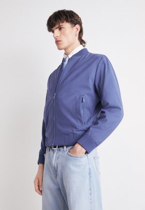 Blue bomber jacket with a zip front, side pockets, ribbed cuffs, and waistband. Paired with light blue jeans and a white shirt.