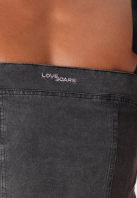 Čierna denimová tkanina, priliehavý strih, s viditeľným šitím; obsahuje vyšívanú nášivku "LOVE SCARS" v ružovej farbe.
