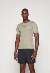 Tričko s krátkým rukávem, přiléhavé, v olivově zelené barvě s černým logem adidas, v kombinaci s černými šortkami s bílými pruhy po stranách.