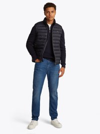 Colete acolchoado azul-marinho com gola alta sobre um suéter azul-marinho, combinado com jeans azul-claro e ténis brancos, apresentando um detalhe de fecho éclair.