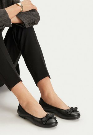 Persona vistiendo pantalones negros ajustados y bailarinas de cuero negro con detalle de lazo, sentada con los brazos cruzados sobre las rodillas.