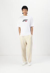White oversized t-shirt with a black graphic and red cherries, paired with beige trousers and white sneakers, standing against a plain background.