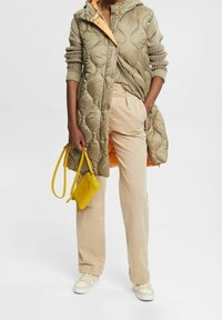 Pikowana kurtka puchowa w oliwkowym zielonym kolorze z kapturem i pomarańczową podszewką, zestawiona z beżowymi spodniami, białymi trampkami oraz małą żółtą torebką.