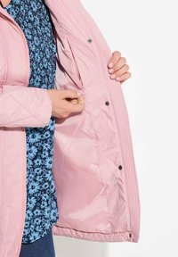 Chaqueta acolchada de color rosa claro con una textura suave, que cuenta con un cierre de botones a presión y un bolsillo interior. Camisa de patrón floral azul visible debajo.