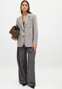 Light grey soft blazer with two front pockets, black buttons, paired with loose dark grey trousers and a textured olive green handbag.