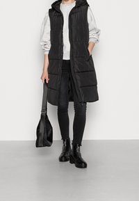 Person som bär en lång svart vadderad ärmlös jacka, grå sweatshirt, svarta smalajeans, svarta stövlar, och håller en svart axelväska.