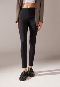 Höga svarta byxor med slimmad passform, tillverkade av stretchigt material, i kombination med svarta loafers och en kort vit topp.