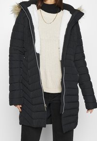 Vadderad svart kappa med huva kantad i konstpäls, dragkedja och innerfoder i fleece. Har sidofickor och midjelång design.