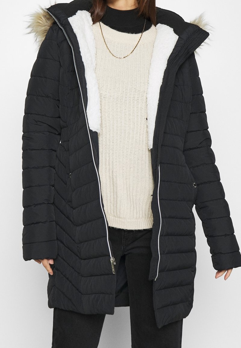 Vadderad svart kappa med huva kantad i konstpäls, dragkedja och innerfoder i fleece. Har sidofickor och midjelång design.