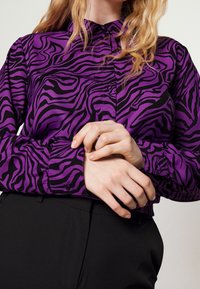 Blouse violette avec des rayures zèbre noires, présentant un col et une fermeture à boutons, complétée par des poignets ajustés. Portée avec un pantalon noir.