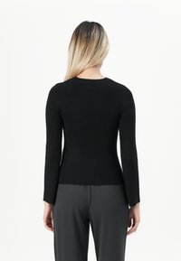 Woman with long blonde hair wearing a fitted black ribbed sweater and dark gray pants, facing away against a plain white background.