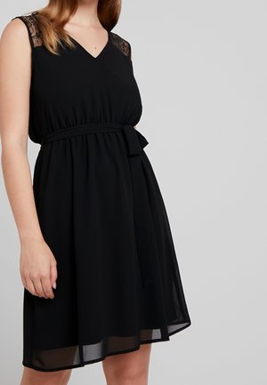 Frau in einem ärmellosen schwarzen Kleid mit Spitzen-Schulterdetails und einem gebundenen Taillengürtel vor einem schlichten hellgrauen Hintergrund.