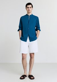 Türkisblaue Hemdbluse mit hohem Kragen, kombiniert mit weißen Shorts. Schwarze Sandalen runden den Look ab. Glattes Material mit lockerer Passform.