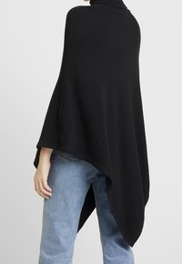 Schwarzer, gestrickter Poncho mit hohem Kragen, asymmetrischem Saum und weiten Ärmeln, kombiniert mit ausgewaschenen blauen Jeans.