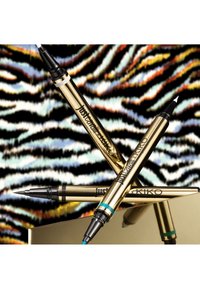 Trois stylos eye-liner liquide dorés avec des pointes noires sur une surface réfléchissante, sur un fond zébré multicolore.