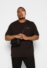 Sort bomulds T-shirt med korte ærmer, der har et lille hvidt Calvin Klein-logo, sammen med en sort skuldertaske med et tekstureret mønster.
