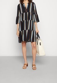 Černé šaty s barevnými svislými pruhy, tříčtvrtečními rukávy a vrstveným designem. Spárovány s hnědými sandály na blokovém podpatku a bílou plážovou taškou.