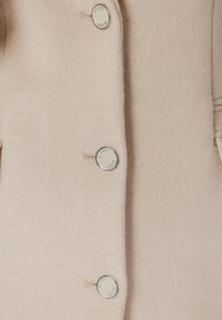 Abrigo de mezcla de lana beige con una textura suave, con botones plateados redondos grabados con "Guess". Diseño entallado y líneas limpias.