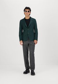 Teksturert mørkegrønn blazer med to knapper, kombinert med grå teksturerte bukser og svarte sko. Modellen har solbriller og en svart skjorte.