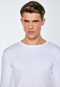 Jauns vīrietis ar īsiem brūniem matiem, valkā vienkāršu baltu garām piedurknēm kreklu, skatās tieši priekšā ar neitrālu izteiksmi uz gaiša fona.