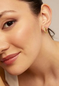 Pendiente de aro dorado con superficie texturizada, que presenta una forma irregular y orgánica. Se lleva en el lóbulo con tonos de piel suaves en el fondo.