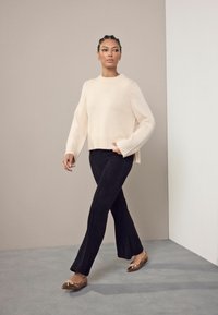 Nainen, jolla on letitetyt hiukset, kävelee päällään kermaisen värinen neule, mustat housut ja ruskeat, kärjet suippoja kenkiä, joissa on nauhakiinnitys, beigeen ja valkoiseen seinää vasten.