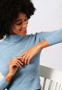 Sølvarmbånd på håndleddet, båret med en lysblå ribbet sweater med høj hals, hvilket viser et tæt på billede af tilbehøret.