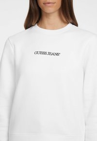Valkoinen huppari, joka on valmistettu sileästä kankaasta. Keskellä musta "GUESS JEANS" -logo. Klassinen pyöreä kaula-aukko ja ribbikuvioidut hihansuut.