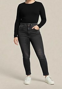 Personne portant un haut à manches longues noir, un jean skinny gris foncé taille haute, des baskets blanches, debout avec une main dans la poche.