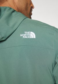 Sudadera verde con una textura suave, que presenta un logo bordado en blanco de The North Face en la parte superior de la espalda y una capucha con cordón.