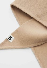 Beiger Fleece-Schal mit weicher Textur, dreieckiger Form und einem kleinen weißen Etikett mit schwarzem Logo am Rand.