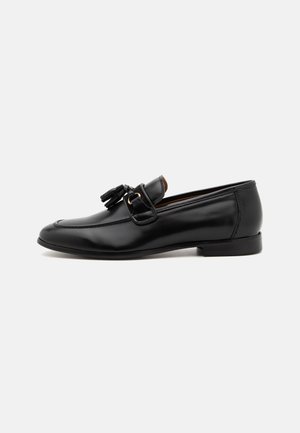 Pantofii din piele neagră pentru bărbați, tip loafer, cu decorare cu ciucuri și toc mic, prezentați în profil pe un fundal alb.