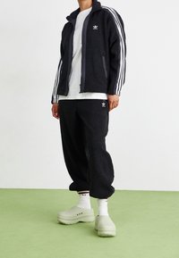 Zwarte fleece trainingspak met wit Adidas-logo en drie witte strepen op elke mouw. Gepaard met lichtgroene klompen en witte sokken.