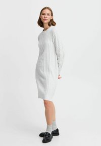 Strickkleid in Hellgrau mit strukturiertem Rautenmuster, langen Ärmeln und großzügigem Schnitt, kombiniert mit schwarzen Loafers und grauen Socken.