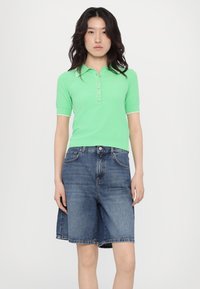 Green knit polo shirt with short sleeves and five buttons; paired with light blue denim shorts that reach mid-thigh.