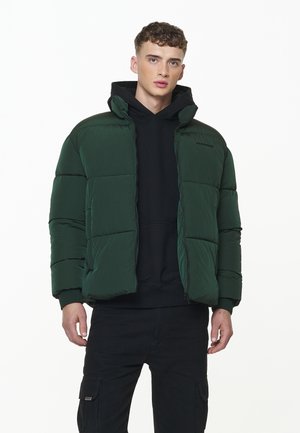 Dark green puffer jacket with a high collar, zip closure, and quilted texture. Worn over a black hoodie, paired with black cargo pants.