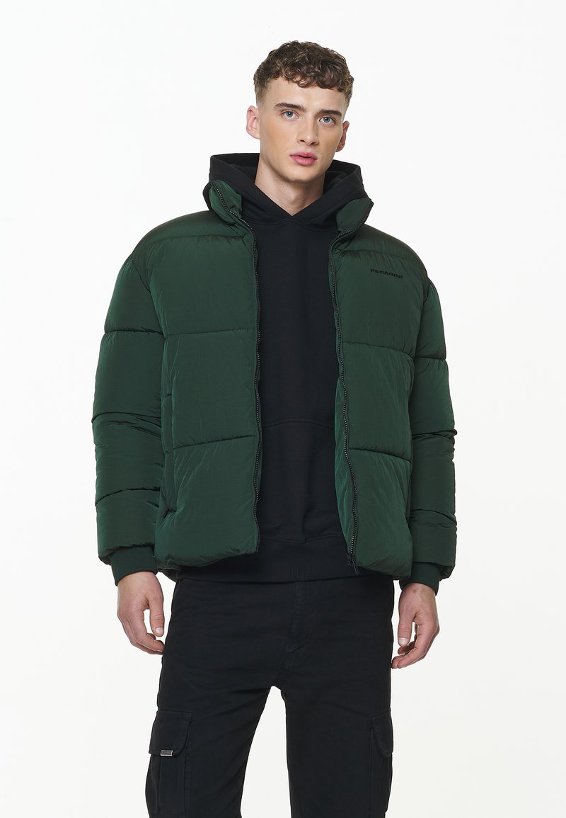 Chaqueta acolchada verde oscura con cuello alto, cierre de cremallera y textura acolchada. Llevada sobre una sudadera negra con capucha, combinada con pantalones cargo negros.